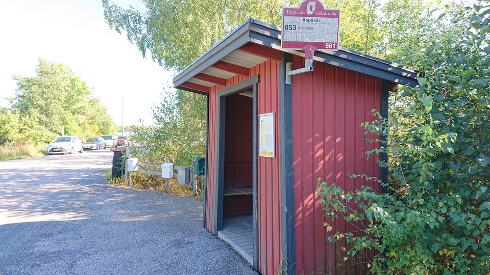 En busskur står vid sidan av vägen. Bakom kuren står brevlådor.Längre bort står flera bilar parkerade vid sidan av vägen.