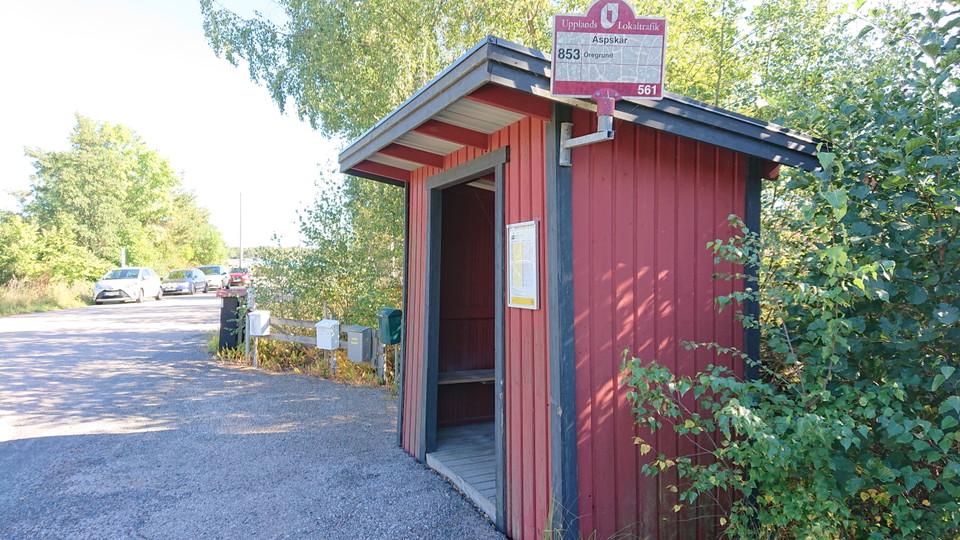 En busskur står vid sidan av vägen. Bakom kuren står brevlådor.Längre bort står flera bilar parkerade vid sidan av vägen.