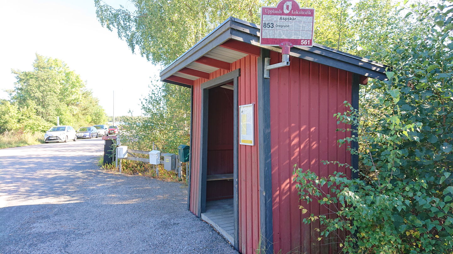 En busskur står vid sidan av vägen. Bakom kuren står brevlådor.Längre bort står flera bilar parkerade vid sidan av vägen.