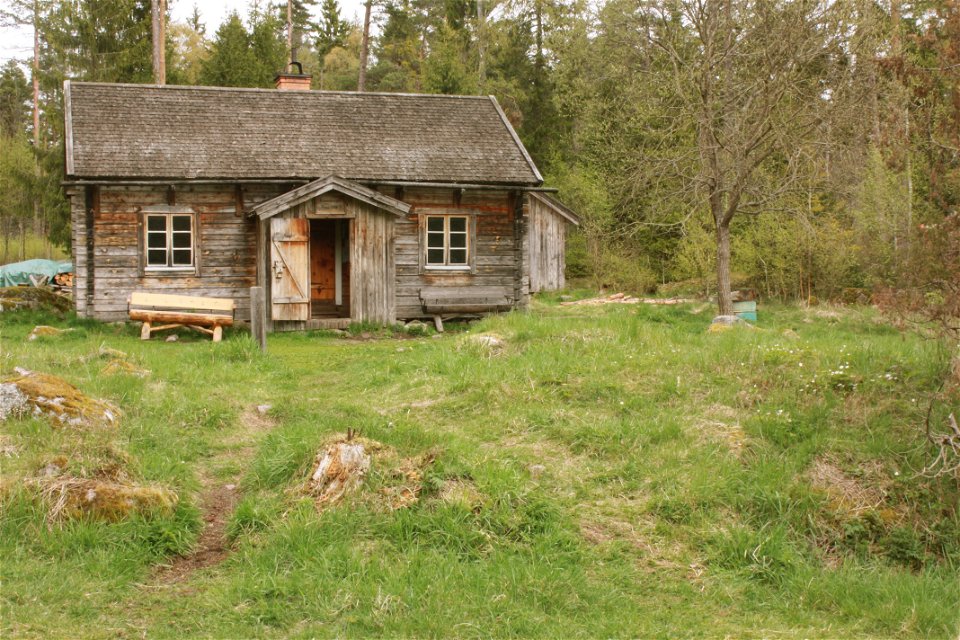 En liten stuga med sittbänk vid ingången står på ojämn gräsmark. Bakom torpet är det tät skog.