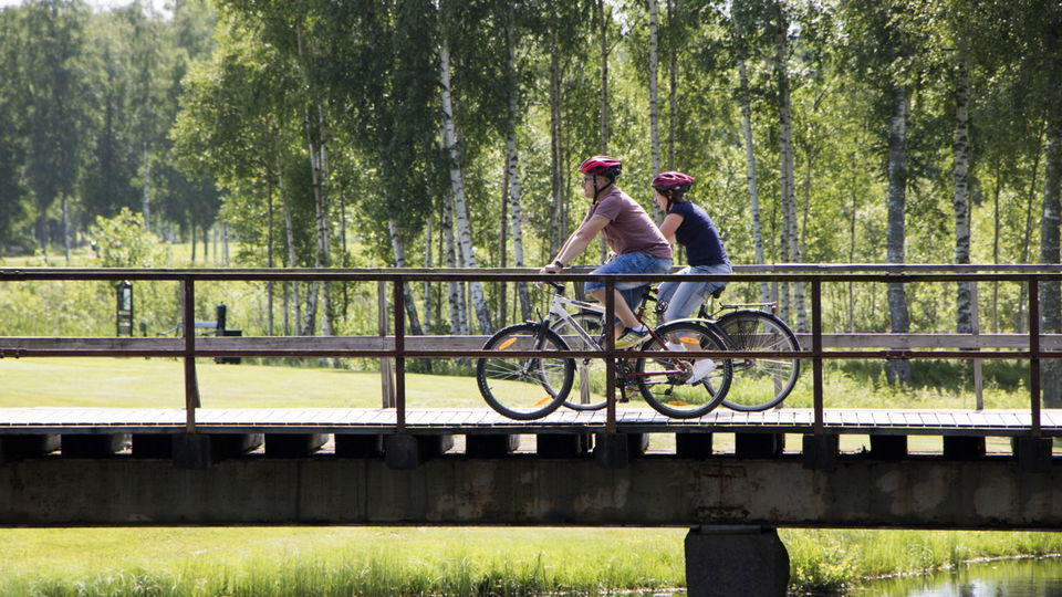 Två personer cyklar över en bro i ett grönskande landskap