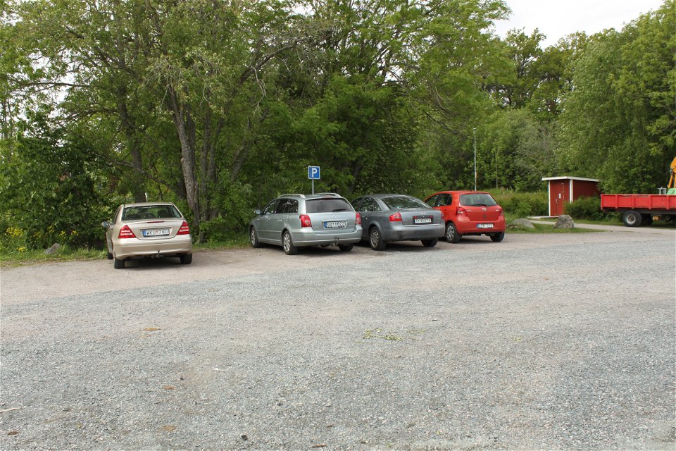 Fyra bilar står bredvid varandra på en stor grusad parkeringsplats. I ena änden av parkeringen står en torrtoalett.