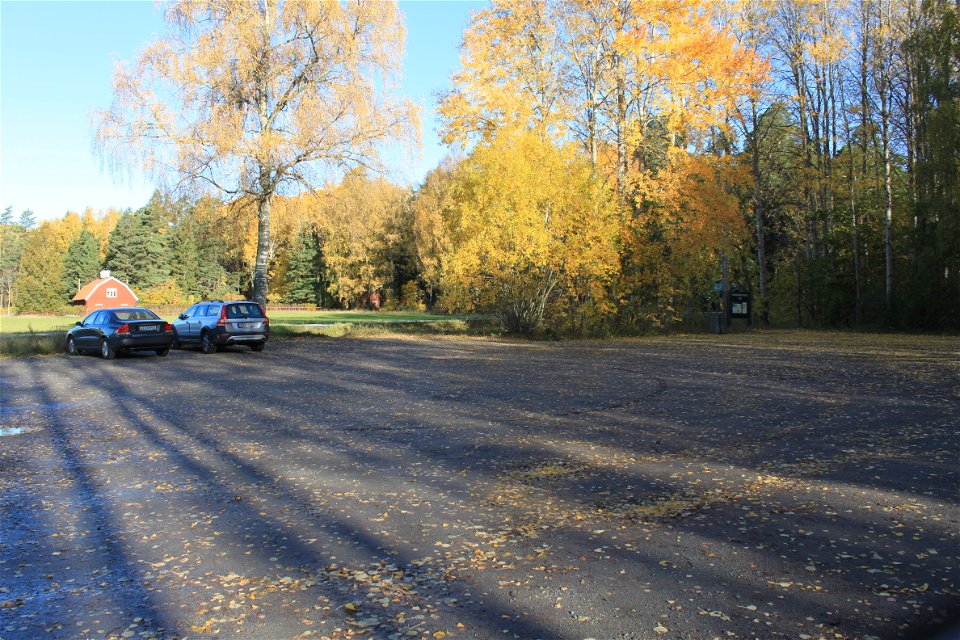Två bilar står på en rymlig grusad parkeringsplats. I bakgrunden är det betesmark, ett hus och skog.