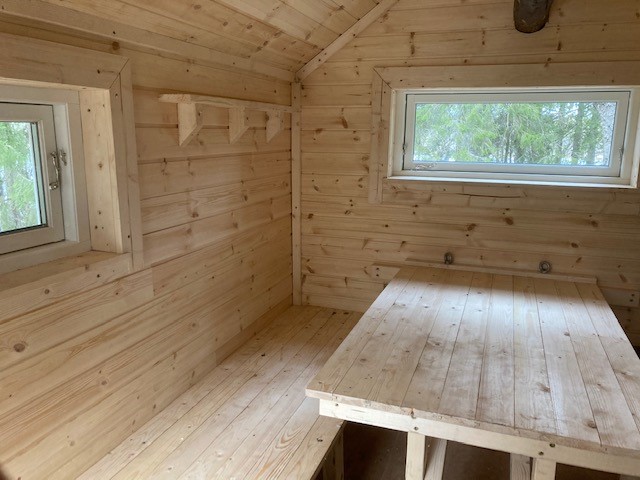 Inuti stuga, väggar och tak i naturfärgat trä, två fönster, ett bord och en bänk i bild.