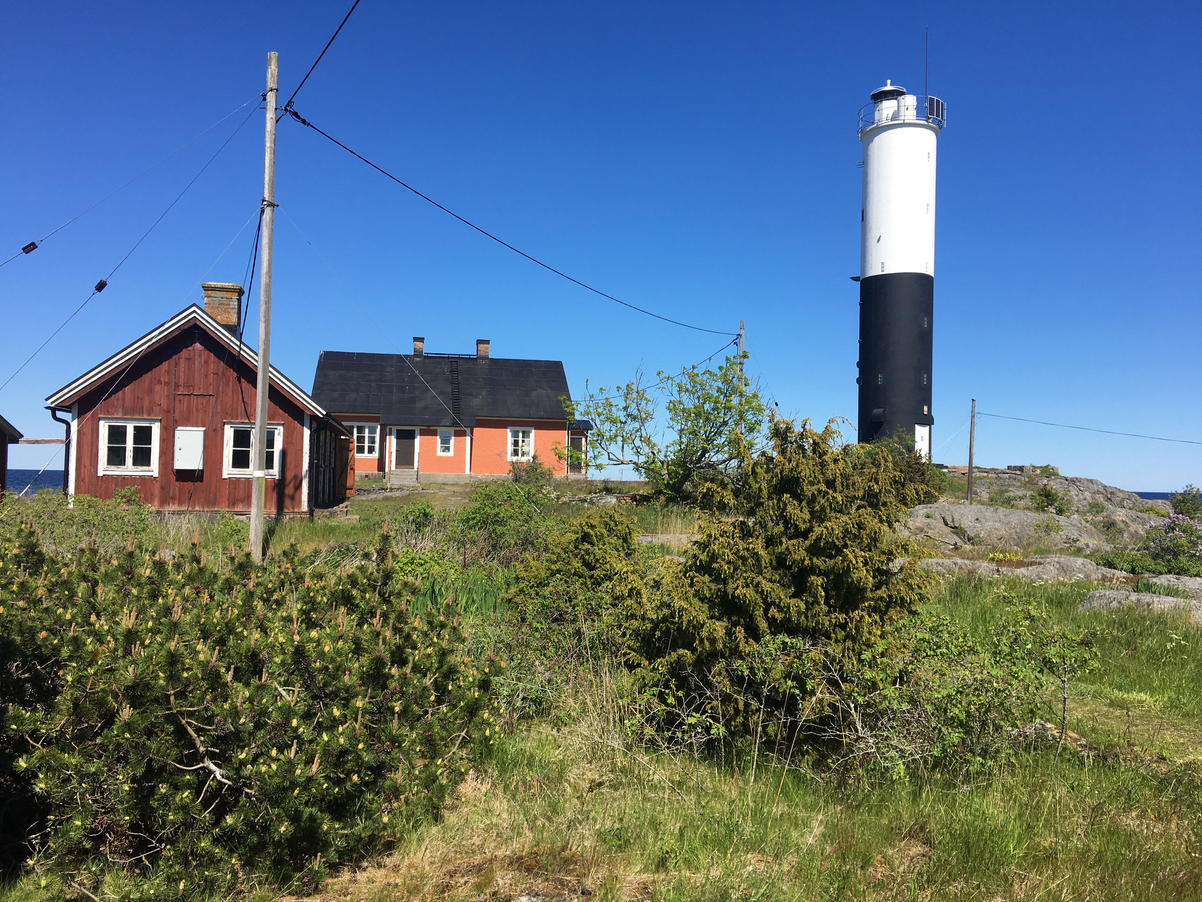 På en ö med ojämn mark och stenhällar står en fyr med två äldre hus vid ena sidan. En hög stolpe med flera elkablar fästa på sig står i närheten av husen.
