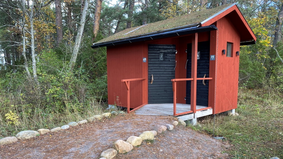 En grusväg med stenar längs kanterna leder fram till en ramp vid en toalettbyggnad. Det finns två dörrar in till toaletten.