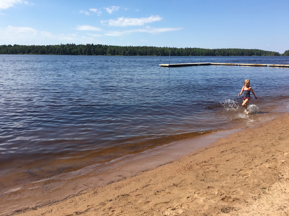 Ett barn leker i strandkanten. En badbrygga ligger i vattnet.