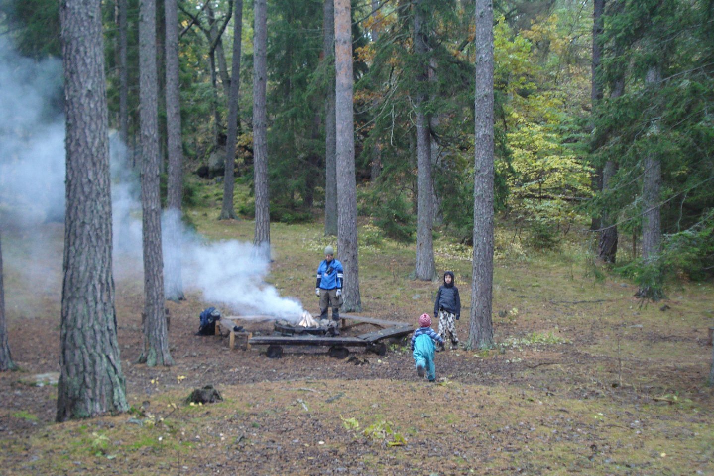 I en skogsglänta står en grillplats med bänkar runtom. Det ryker kraftigt från elden på grillplatsen och tre barn tittar på.