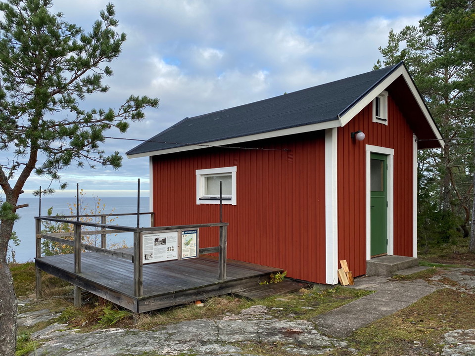 En liten stuga med altan står på en klippa vid havet. Altanen har ett högt räcke med en informationsskylt.