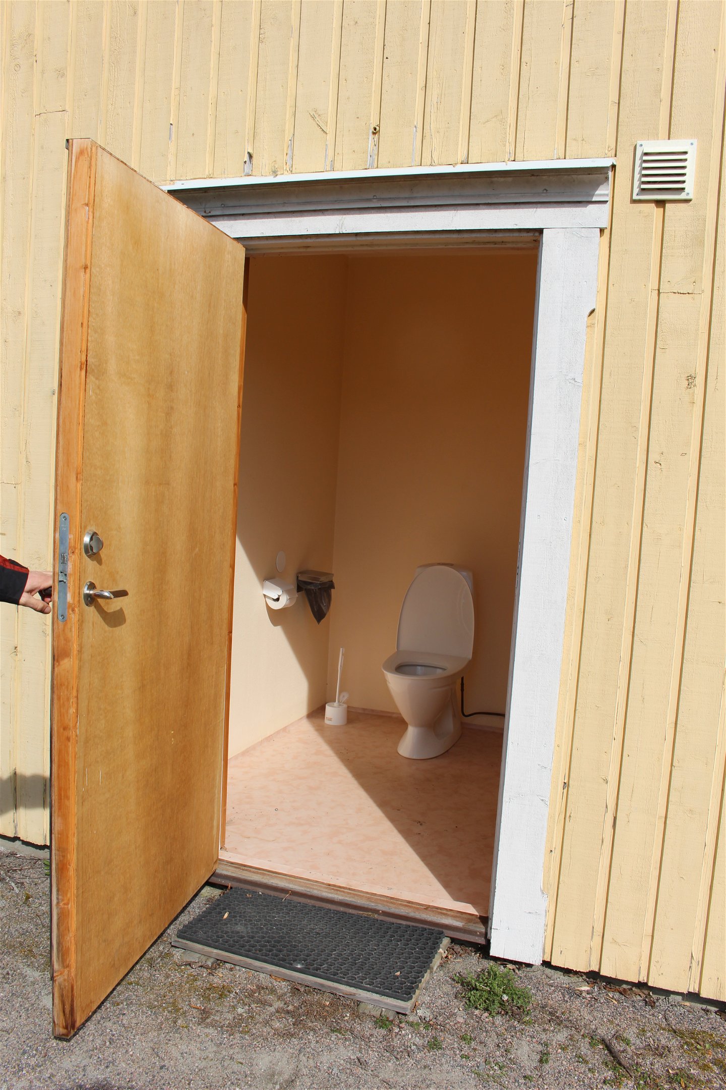 En person håller upp dörren in till en toalett. Inne på toaletten står en toastol, toapapper och en toaborste.