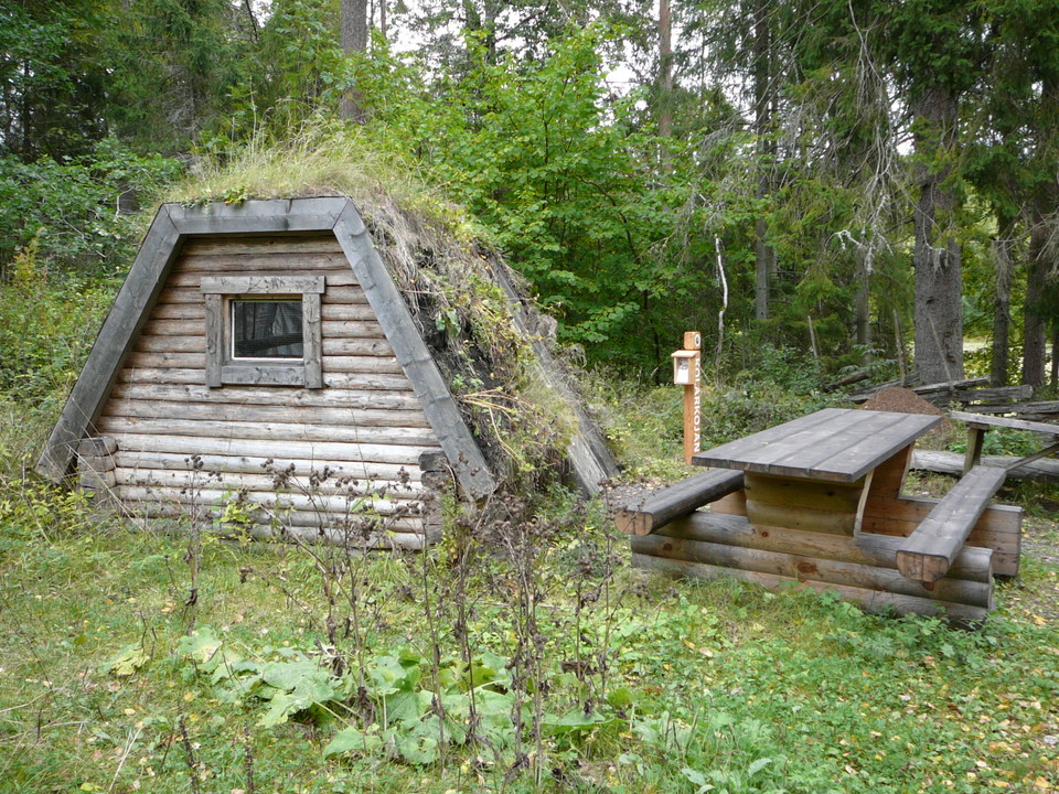 I skogen står en koja med ett litet fönster, det växer mossa på taket. Vid kojan står ett bänkbord och en stolpe med broschyrer.