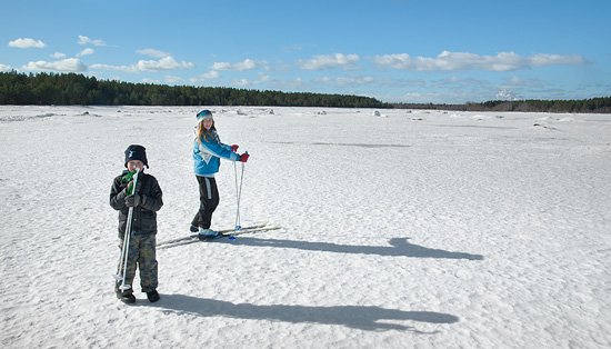 Två barn står på en jämn snötäckt yta. Ett av barnen står på skidor och det andra barnet håller i ett par skidstavar.