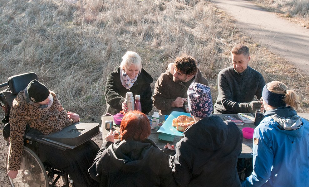 Sju personer sitter och fikar runt ett bord i naturen.