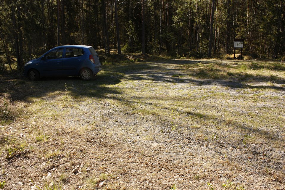 En bil står på en stor parkeringsplats med en informationstavla. Parkeringen är omringad av skog och underlaget är ojämt med jord och gräs.