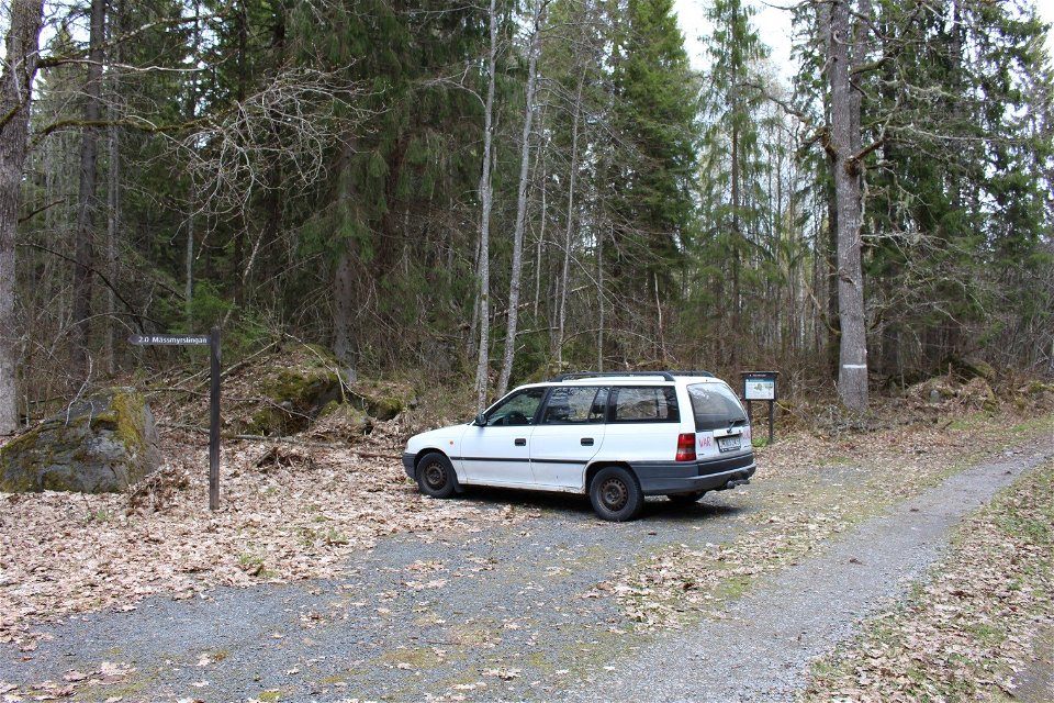 En bil står på en liten grusad parkeringsplats vid en skog. På parkeringen finns en informationsskylt och en vägvisare.