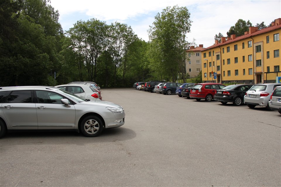 På en asfalterad parkering står många bilar parkerade bredvid varandra. På ena sidan parkeringen är det skog och på andra sidan är det bostadshus.
