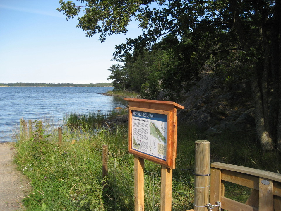 En informationsskylt står på en strand vid havet. Bakom skylten är det skog och klippor.