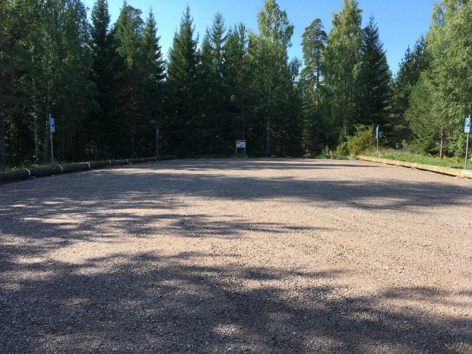 En mycket rymlig grusad parkeringsplats med liggande stockar runt kanterna. I bakgrunden är det skog.