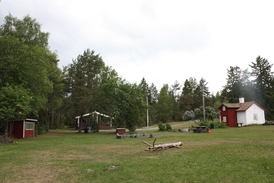 Tre stugor med en stor gräsyta omringad av lövträd. Flera bänkar och bord står på gräset.