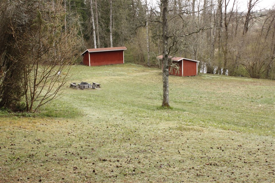 På en öppen gräsyta som sluttar lätt neråt står en grillplats och två mindre bodar.