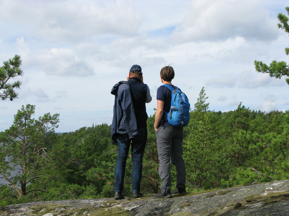 En man och en kvinna står uppe på en höjd med ryggen mot bilden. Nedanför dem står många tallar och man har utsikt över vattnet.