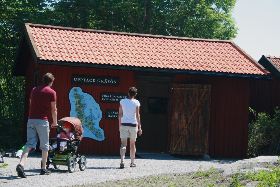 Två vuxna går längst en grusväg mot en öppen bod. En av vuxna rullar ett barnvag med ett barn i. 