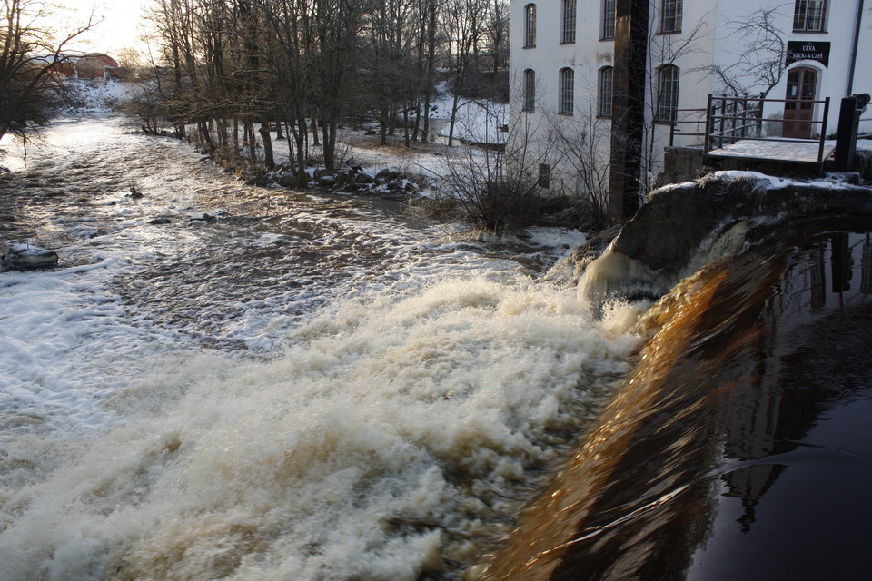 En fors med snabbt strömmande vatten. Vid vattnet står en stor byggnad med många fönster.