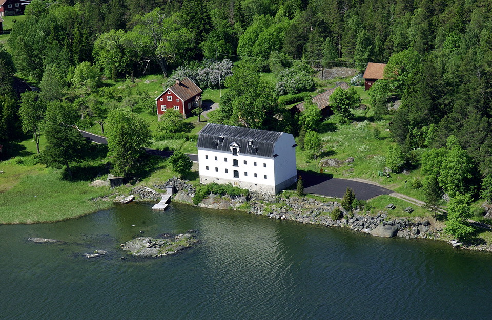I en havsvik står ett stort järnmagasin intill vattnet. Bredvid finns en parkeringsplats och en liten brygga ut till vattnet.