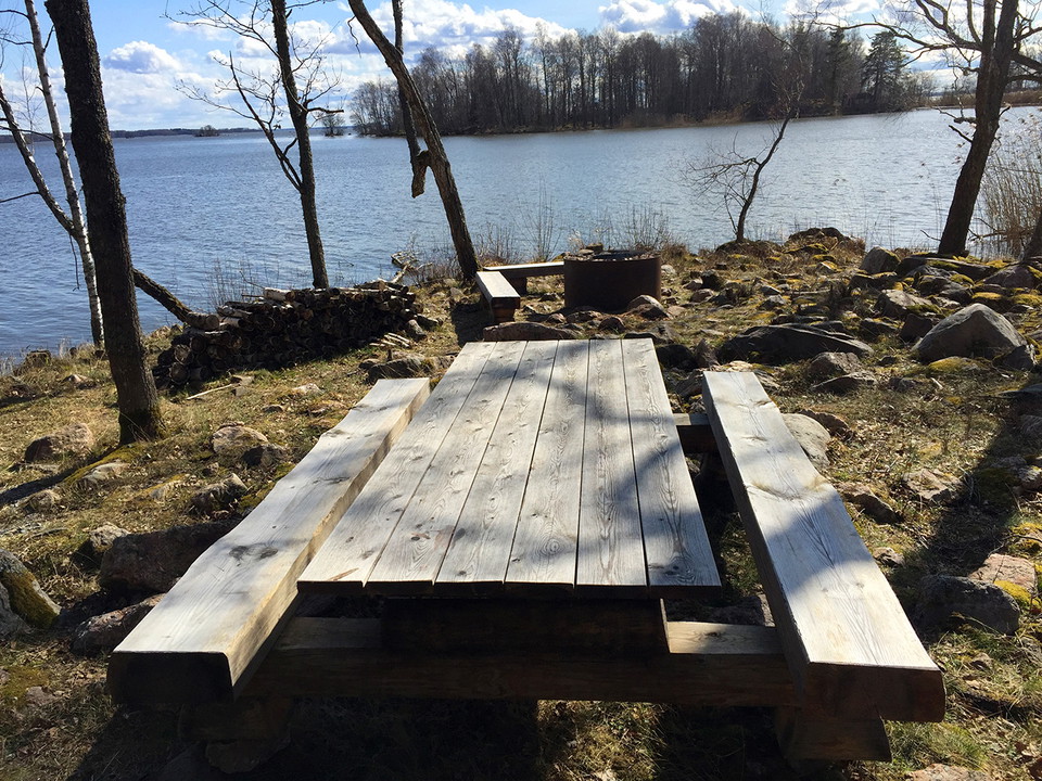 Träbord, träbänkar och eldstad. Strandkant och vatten i bakgrunden. Vårsolen skiner.