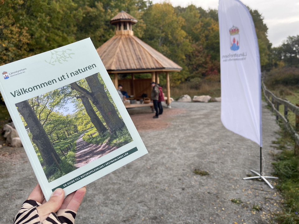 En bok med texten "Välkommen ut i naturen" hålls upp framför ett vindskydd där det står fyra män.