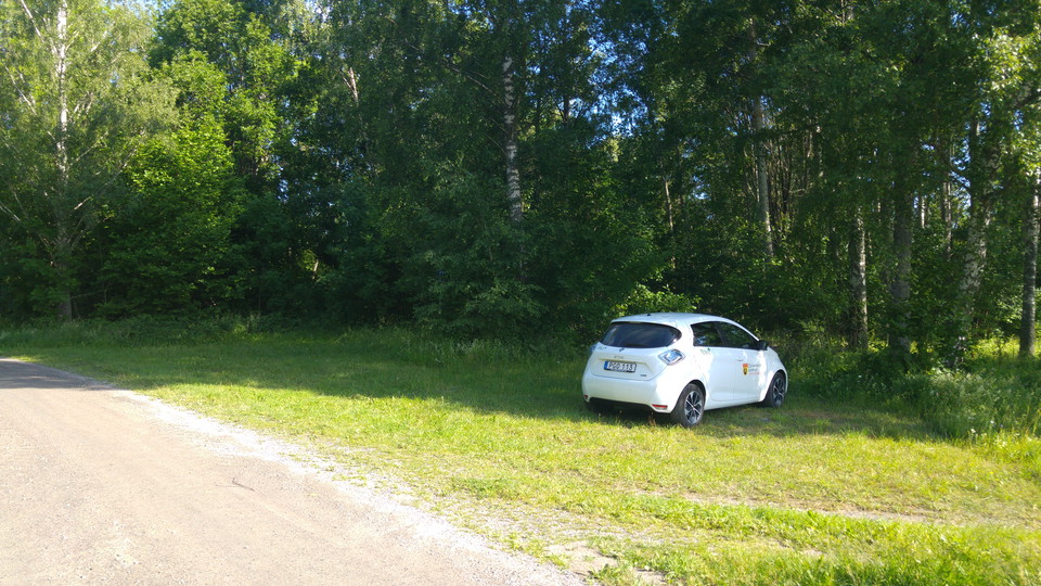 En bil står på en mindre parkeringsplats vid en väg. Underlaget på parkeringen är gräs.