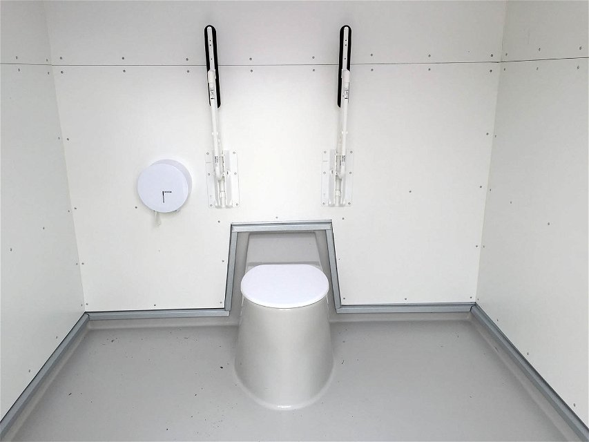 Toalettstol med nerfällt lock. Två armstöd uppfälda mot väggen. En toalettpappershållare till vänster om toalettstolen.