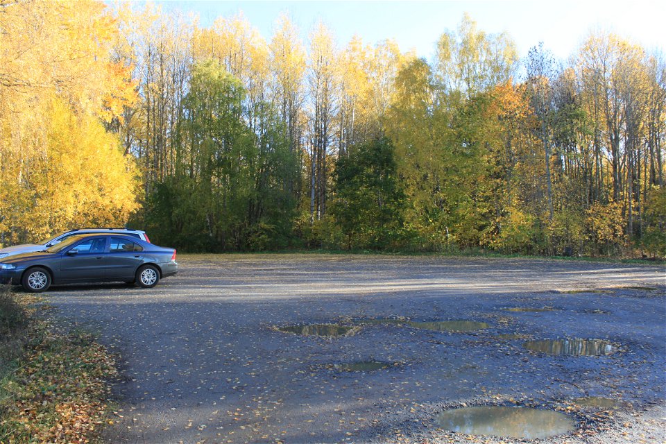 Två bilar står på en stor grusad parkering med skog runtom. Det finns flera vattenfyllda gropar på parkeringen.