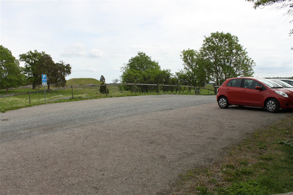 På en bred grusad parkeringsplats står bilar parkerade. I bakgrunden finns en hage och en hög kulle.