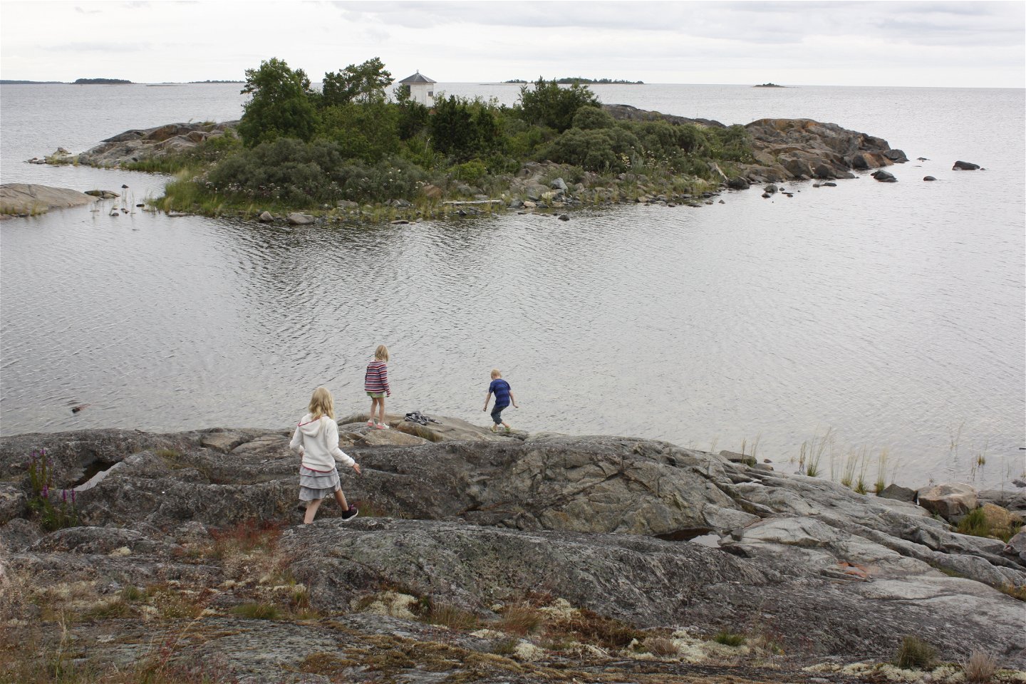 Tre barn går på en klipphäll vid havet. I bakgrunden finns en liten ö med många buskar.
