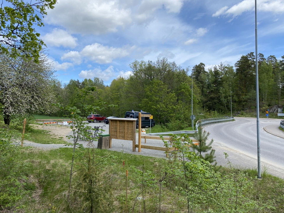 Vid en bilväg finns en parkering med plats för flera bilar. På kanten av parkeringen finns en soptunna och en informationstavla.