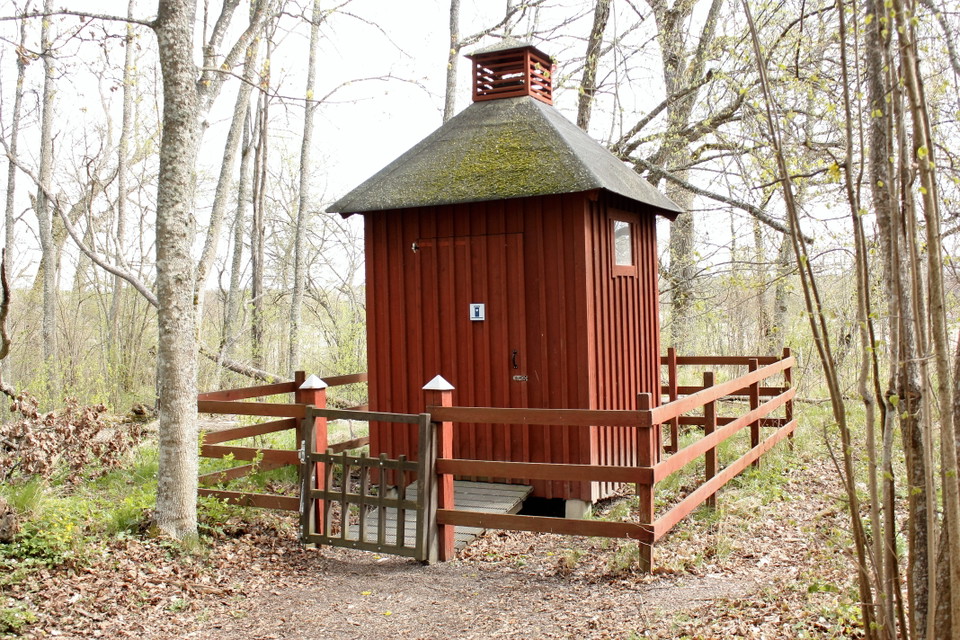 En toalettbyggnad med staket runtom. En grind och en liten spång leder fram till toalettdörren.