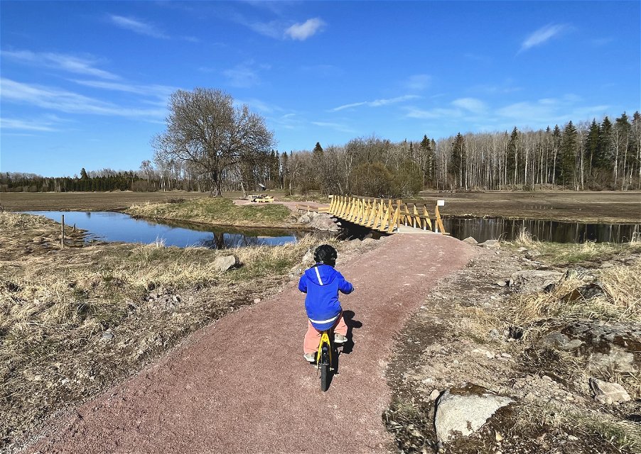 Ett litet barn med blå jacka cyklar på en smal grusväg som leder till en bro. Bron går över ett vattendrag, på andra sidan finns en rastplats, åkrar och i bakgrunden en skogsridå.