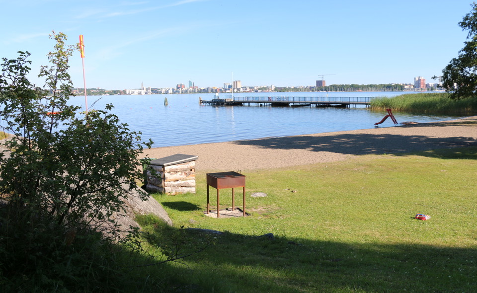 Sandstrand med grill, rutschkana och en lång brygga. I bakgrunden skymtas Västerås.