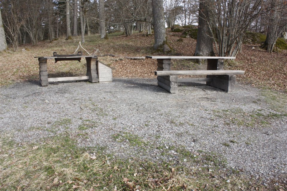 På en grusplan står ett bänkbord och ett grillbord. I bakgrunden är det skog.