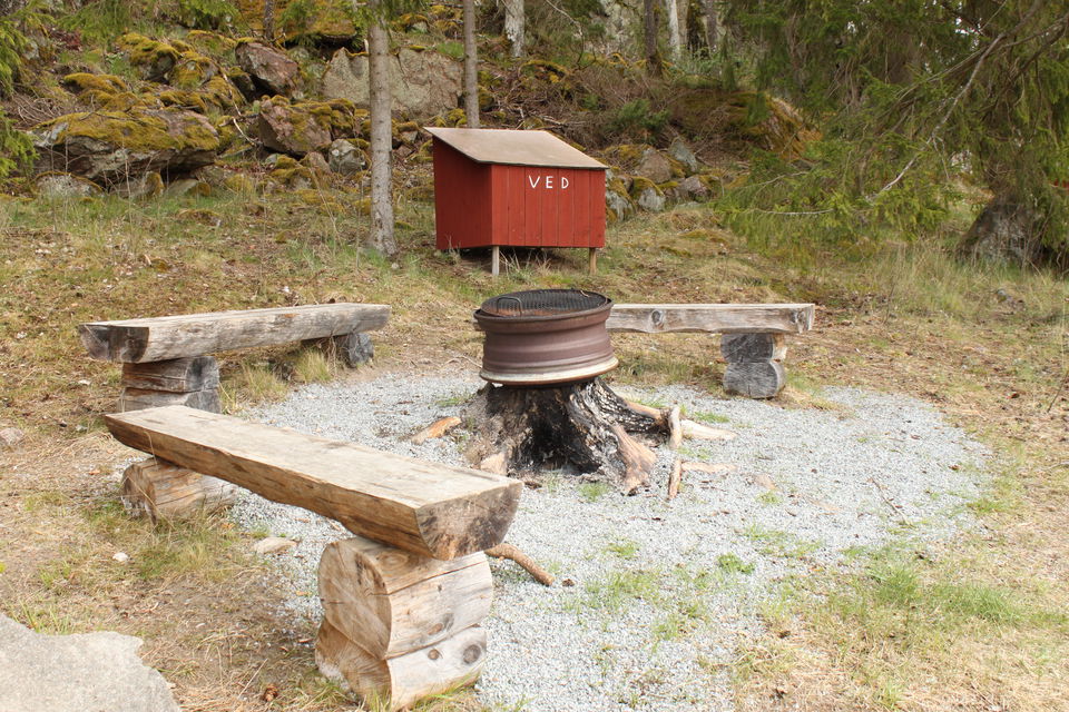 En grusad grillplats med sittbänkar och ett litet vedförråd en bit bort.