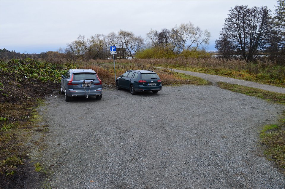 En liten grusad parkeringsplats med två bilar på. Det är nära till gångvägen som löper längs med Fyrisån.