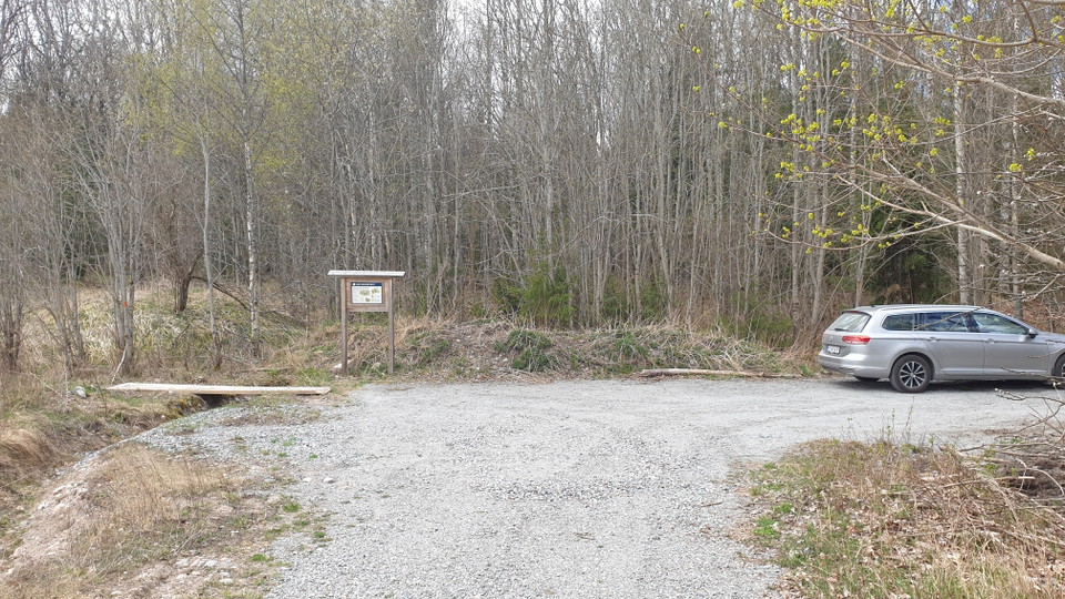 En grusväg i en skog, en parkeringsficka till höger där det står en grå bil parkerad.