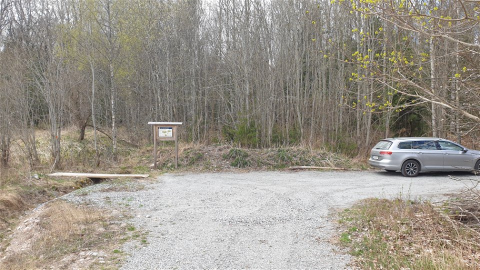En grusväg i en skog, en parkeringsficka till höger där det står en grå bil parkerad.