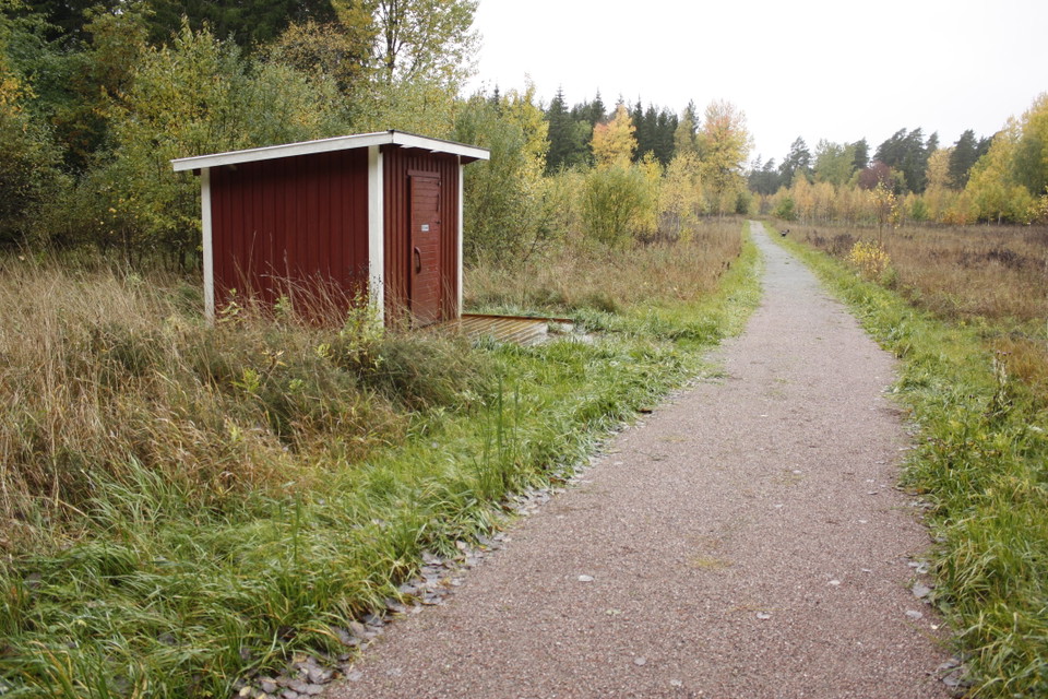 En bred grusväg med skog runtom. Vid kanten av vägen står en torrtoalett med en ramp framför.