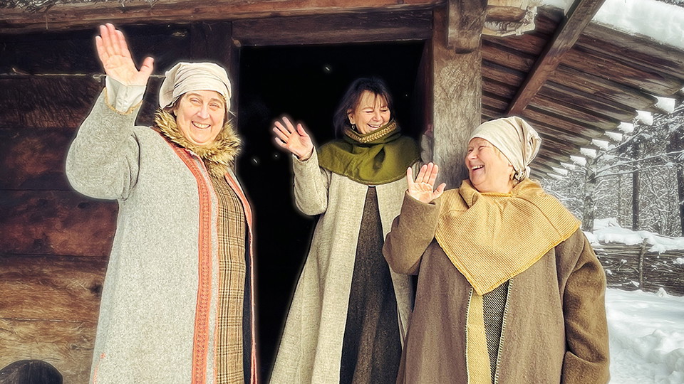 Tre kvinnor i historiska kläder ler och vinkar mot kameran. De står utanför ett trähus.