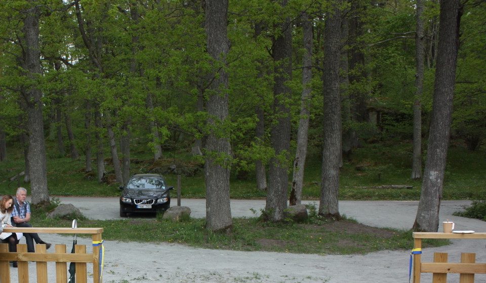En bil står på en grusad parkeringsplats. Mitt på parkeringen finns en liten dunge med några träd. En person sitter på en bänk vid sidan av parkeringen.