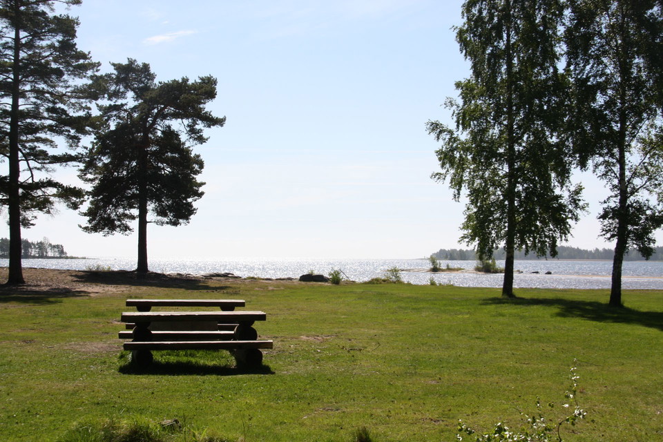 Badplats i bakgrunden, en gräsplätt i framsidan med fikabord och bänkar