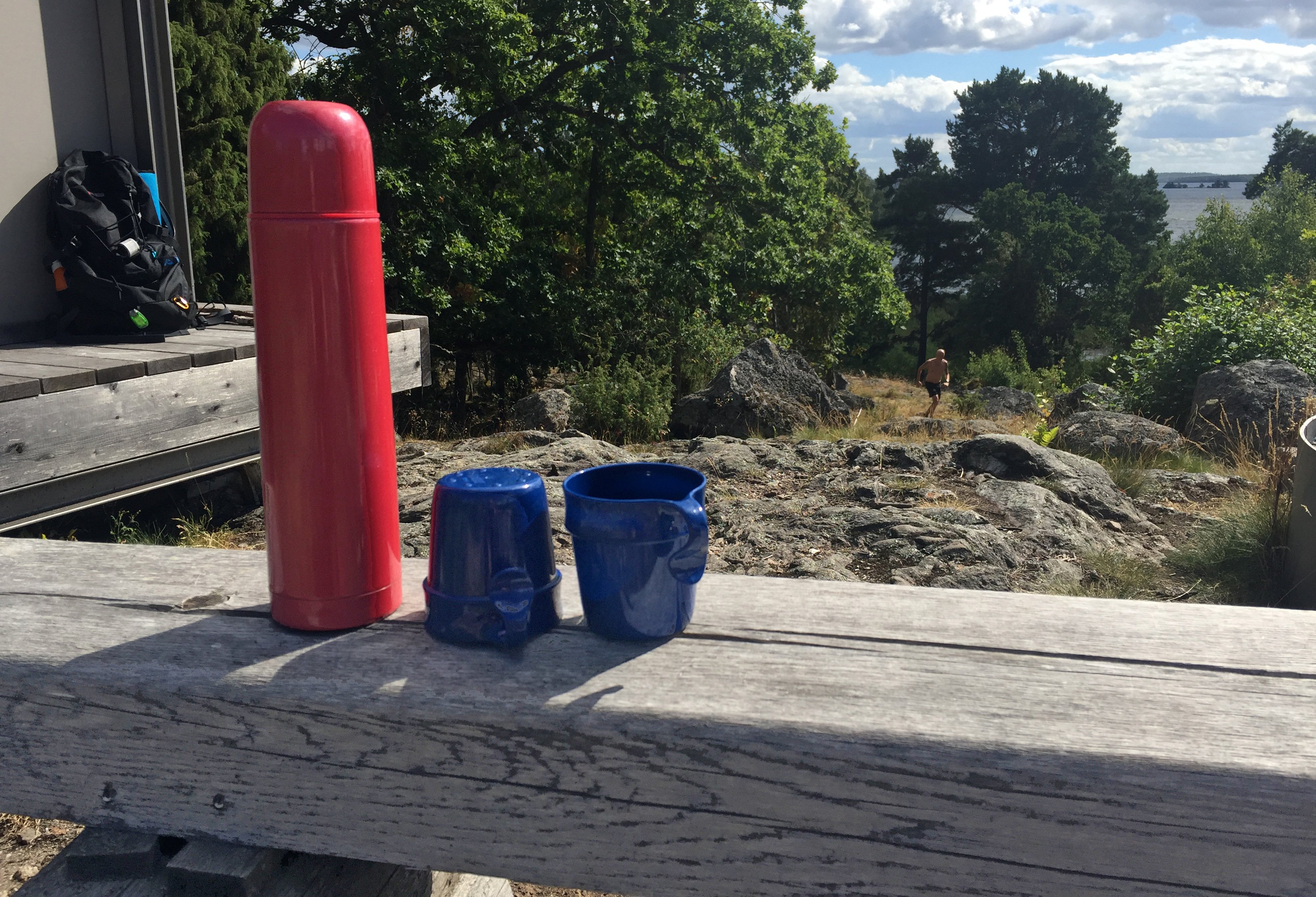 Röd termos och två blå muggar på träbänk.