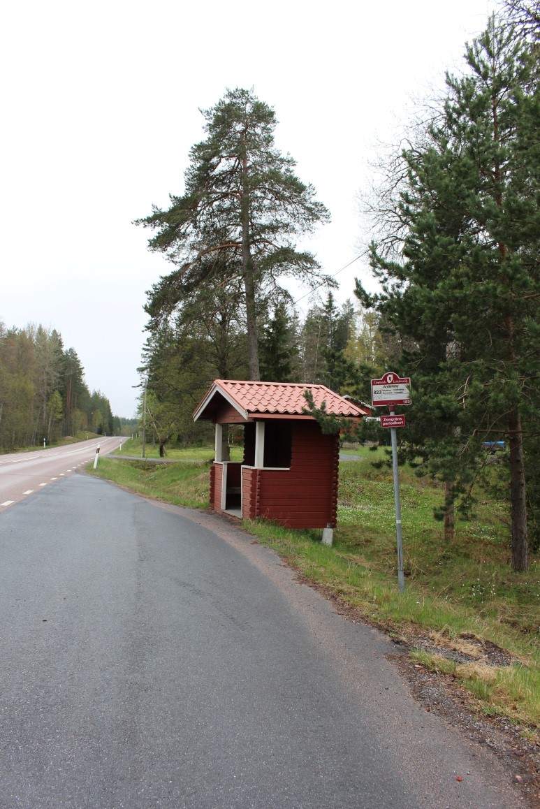På en busshålssplats vid en väg står en busskur och en hållplatsskylt. Busskuren är av trä med lågt tak.